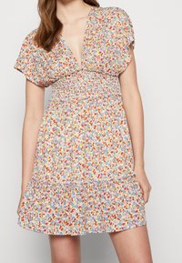 Robe fleurie avec un décolleté en V profond, manches courtes bouffantes, taille smockée et ourlet à volants. Présente de petites fleurs colorées sur un fond clair.