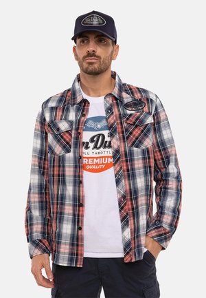 Homme portant une casquette bleu marine, un t-shirt blanc avec un motif graphique, une chemise à carreaux rouge et bleue boutonnée, et un pantalon cargo sombre, debout avec une main dans la poche.