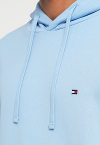 Sweat à capuche bleu clair avec cordons et petit logo rouge, blanc et marine brodé sur la poitrine.