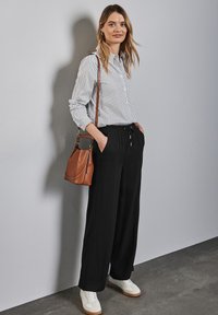 Chemise rayée, pantalon noir ample, baskets blanches avec des accents beige. Sac bandoulière marron. Le modèle se tient contre un mur gris.