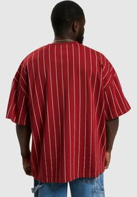 Camicia in cotone oversize a righe bordeaux con maniche corte, caratterizzata da strisce verticali bianche e una vestibilità morbida. La vista posteriore mette in evidenza il design.