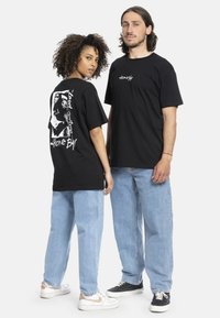 Sorte t-skjorter med grafisk design på baksiden, fet tekst og avslappet passform. Kombinert med lyseblå jeans og joggesko.