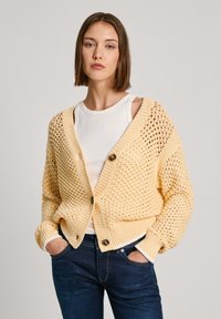 Žlutý pletený kardigan s volným, otevřeným vzorem. Má výstřih do V, dva velké knoflíky a žebrované manžety. Nosený přes bílý top bez rukávů.