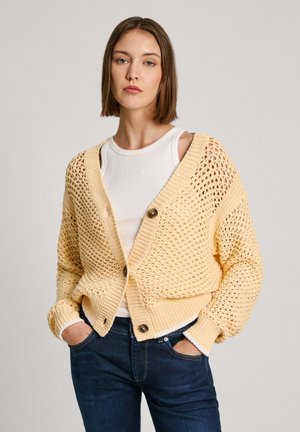 Cardigan giallo in maglia con trama aperta e morbida. Presenta un collo a V, due grandi bottoni e polsini a coste. Indossato sopra un top bianco.