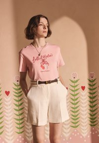 Růžové bavlněné tričko s červeným květinovým motivem a nápisem, doplněné béžovými lněnými šortkami, zdobené hnědým opaskem.