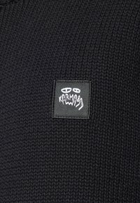 Black knitted sweater featuring a square black label with white graphic elements and text, showcasing a textured material and simple design.