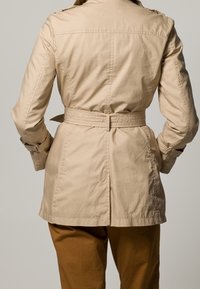 Beige trench coat med bältesmidja, knäppte ärmslut och axelapplikationer; har ett slätt, strukturerat tyg.