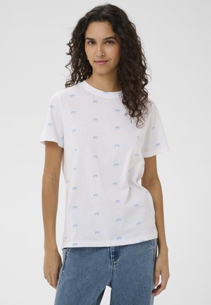 Žena s kučeravými vlasmi, oblečená v bielom tričku so vzorom malých modrých mašličiek a modrých džínsach, stojaca pred jednoduchým pozadím.