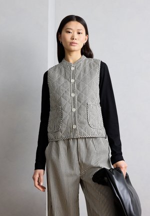 Jeune femme portant un haut à manches longues noires avec un gilet et un pantalon à rayures verticales noir et blanc, tenant un sac en cuir noir à ses côtés.