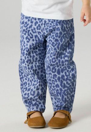 Dieťa oblečené v modrých nohaviciach s leopardím vzorom a hnedých topánkach s remienkami stojí na svetlošedom pozadí.