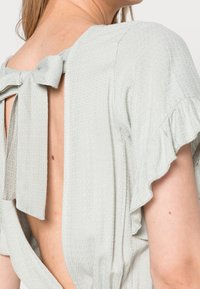 Světlounce zelená halenka s nadýchanými rukávy, texturovaným vzorem a mašlí na zádech, s hlubokým otevřeným zadním dílem.