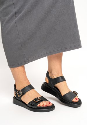 Füße in schwarzen Ledersandalen mit Schnallen und rotem Nagellack, kombiniert mit einem grauen, gerippten Rock, der unterhalb der Knie endet.