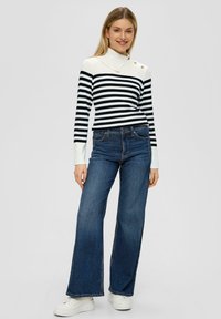 Navy- und weiß gestreifter Strickpullover mit hohem Kragen und Knopfakzenten, kombiniert mit weit geschnittenen blauen Jeans und weißen Sneakers.