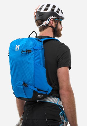 Sac à dos en millet bleu avec une texture hexagonale, des bretelles réglables et des accents noirs. Porté par une personne avec un casque et un équipement d'escalade.