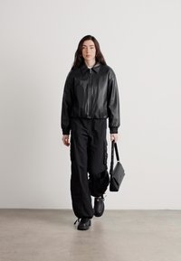 Giacca bomber in ecopelle nera con chiusura a zip, abbinata a pantaloni cargo neri larghi e scarpe nere robuste, mentre tiene in mano una piccola borsa a tracolla.