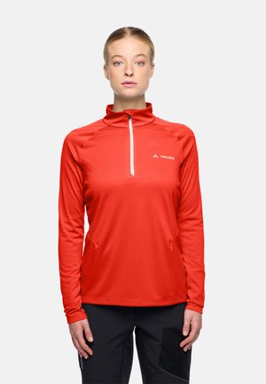 Junge Frau steht, trägt ein rotes Langarm-Sportoberteil mit Reißverschluss und schwarze Hosen, vor einem schlichten weißen Hintergrund.