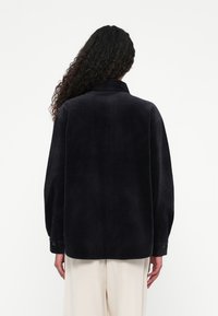 Person med langt, krøllet hår iført sort kordfløjljakke og lys beige bukser, vist fra ryggen mod en ensfarvet hvid baggrund.