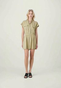 Χακί κοντός φορμαρισμένος σορτς με κοντά μανίκια, μπροστινό φερμουάρ, ελαστική μέση και δύο τσέπες στο στήθος. Το μοντέλο φορά μαύρες παντόφλες.