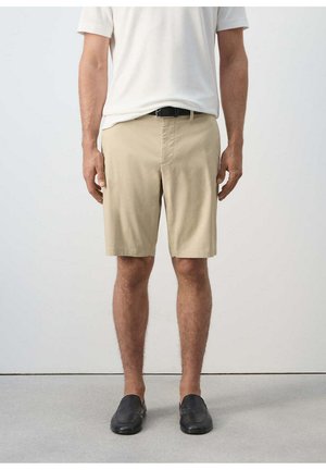 Hombre que lleva pantalones cortos beige, cinturón negro, camisa blanca de manga corta y zapatos negros sin cordones, de pie frente a un fondo claro y liso.