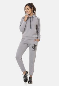 Grauer Kapuzenpullover mit Kordelzug, versehen mit einem Logo auf der Vorderseite. Kombiniert mit passenden Jogginghosen mit gesticktem Logo und Bündchen an den Knöcheln.