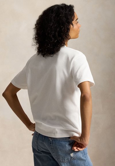 Kobieta o kręconych włosach, ubrana w prostą białą koszulkę z krótkim rękawem i niebieskie dżinsy, stojąca z rękami na biodrach, zwrócona bokiem na neutralnym tle.