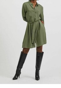 Olivgrön skjortklänning med knäppning framtill och midjebälte. Bärs med knähöga, svarta stövlar med krokodilmönster. Slät tygstruktur.