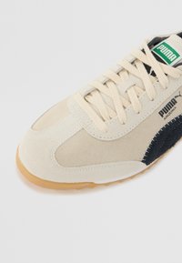 Beige und marineblaue Wildledersneaker mit einer Gummisohle, runder Zehenpartie, Schnürverschluss und auffälligem Puma-Logo auf der Zunge und der Seite.