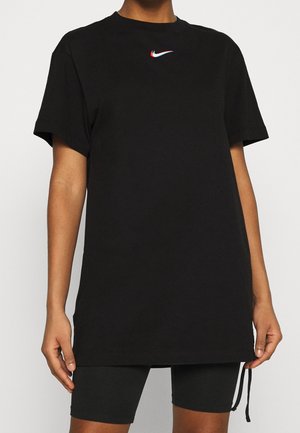 Personne portant un t-shirt Nike noir ample à manches courtes avec un petit logo swoosh multicolore sur la poitrine, associé à un short noir.
