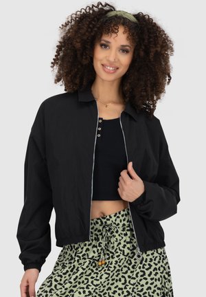 Schwarze, verkürzte Bomberjacke mit Frontreißverschluss, langen Ärmeln und elastischen Bündchen, getragen über einem schwarzen Crop-Top, kombiniert mit Leopardenmuster-Hosen.