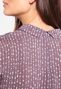 Damskjorta med krage, korta ärmar och lila tyg med vita prickiga mönster, som visar en strukturerad yta.