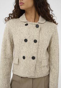 Bežani pleteni sako s dvostrukim kopčanjem, crnim gumbima, ovratnikom s udubljenjima i dva prednja džepa. Teksturirana tkanina i skraćeni kroj.