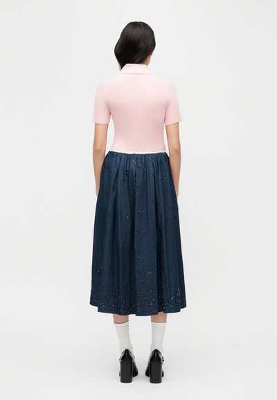 Γυναίκα που βλέπει προς τα πίσω φορώντας ροζ κοντομάνικη μπλούζα και μακριά σκούρα μπλε φούστα με λεπτομέρειες από παγιέτες σε σχήμα λουλουδιών, άσπρες κάλτσες και μαύρες γόβες.