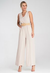 Beżowy kombinezon bez rękawów z dekoltem w kształcie V, szerokimi nogawkami i plecionym paskiem. Miękka tkanina, dobrze się układa. Połączony z przezroczystymi sandałami na obcasie.