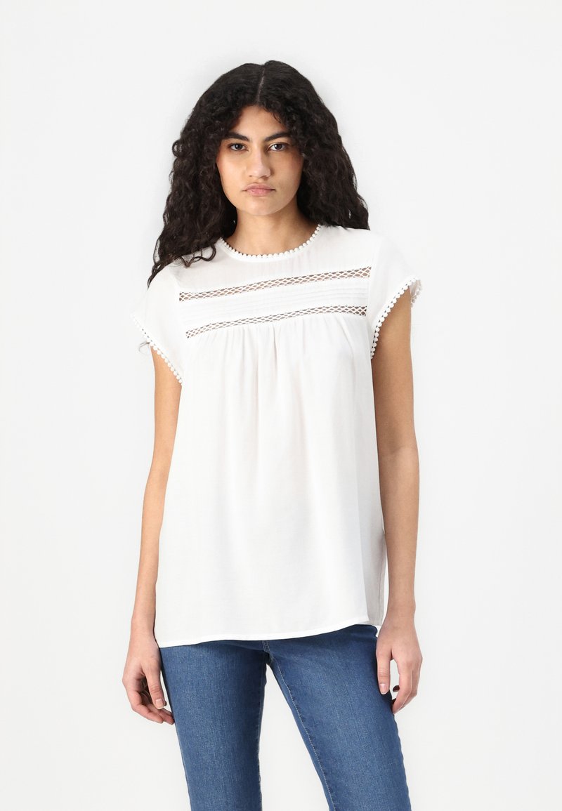 Blusa blanca de manga corta con detalles de encaje en el pecho y un escote redondeado. Diseño fluido con acentos texturizados en las mangas.