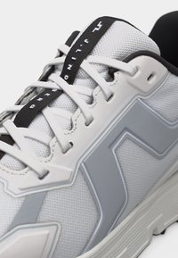 Κοντινό πλάνο λευκού και γκρι αθλητικού παπουτσιού με λευκά κορδόνια και μαύρη θηλιά τράβηγμα με λευκό κείμενο, που δείχνει υλικά πλέγματος και καουτσούκ.