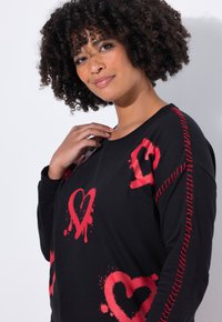Zwarte sweatshirt met rode hartontwerpen en spettereffecten. Contrasterende stiksels langs de schouders voegen details toe. Zachte, gestructureerde stof.