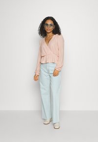 Top envolvente rosa claro com padrão texturizado, mangas longas e decote em V, combinado com umas calças largas de cintura alta, em azul claro, e sapatos beges.