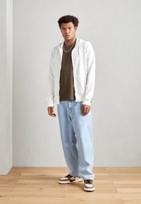 Bijela zip-up hoodie preko maslinaste majice, u kombinaciji s svijetlo plavim širokim trapericama i tenisicama s smeškastim detaljima na drvenom podu.