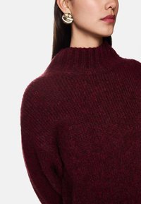 Bordeaux gebreide trui met een hoge, geribbelde kraag. Gouden ronde oorbellen met een glanzende afwerking complementeren de look. De textuur voelt zacht en warm aan.