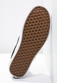 Semelle d'une chaussure de sport avec une semelle en caoutchouc marron présentant un motif en losange, bordée de matériau blanc et d'une tige textile noire.