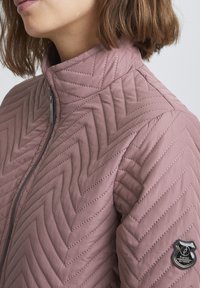 Prošívaná bunda v jemné mauve barvě s vzorem chevron. Má vysoký límec a nášivku na levém rukávu. Zapínání na zip.