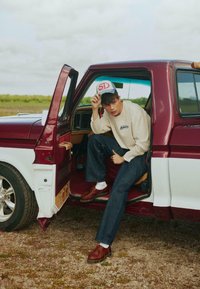 Jeune homme portant des vêtements décontractés et une casquette assis dans la porte ouverte d'un pick-up vintage rouge et blanc sur du gravier.