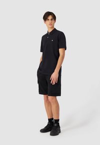 Svart polo skjorte med korte ermer og krage, kombinert med svarte cargo shorts med lommer. Barbert med svarte sokker og sneakers.