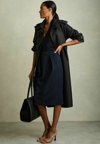 Cappotto nero, gonna blu scuro aderente con le pieghe, décolleté beige con le punte scoperte e una borsa nera. Texture lisce, design slanciato.