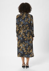 Robe longue noire avec un motif floral en or et bleu. Elle présente des manches longues et une ceinture à la taille, avec une silhouette fluide et un ourlet fendu.