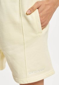 Lys gul bomullsshortser med en sidelomme, med en sømmet kant og "PEGADOR" brodert i hvitt på den nederste delen av benet.