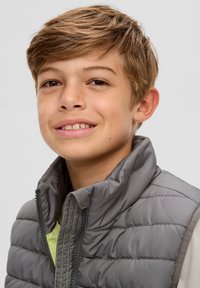 Grau gepolsterte Weste mit hohem Kragen und grünen Nähten, über einem hellgrünen Hemd getragen. Verfügt über einen Frontreißverschluss und eine gesteppten Optik.