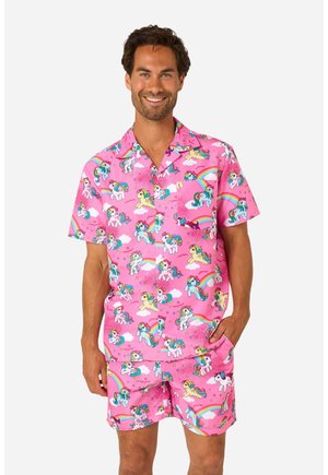 Hombre con camisa rosa de manga corta y pantalones cortos con dibujos coloridos de unicornios y arcoíris, sonriendo con una mano en el bolsillo.