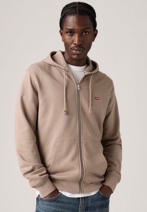 Beige zip-up hoodie with drawstrings, front pockets, and a small red logo. Worn over a white shirt, made from soft, textured fabric.