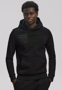 Fekete kapucnis pulóver kenguruzsebbel, szürkével nyomtatva "CAMP DAVID" és "SPORTS CLUB" felirattal. Simaságával, állítható kapucnijával és egyszerű dizájnjával tűnik ki.